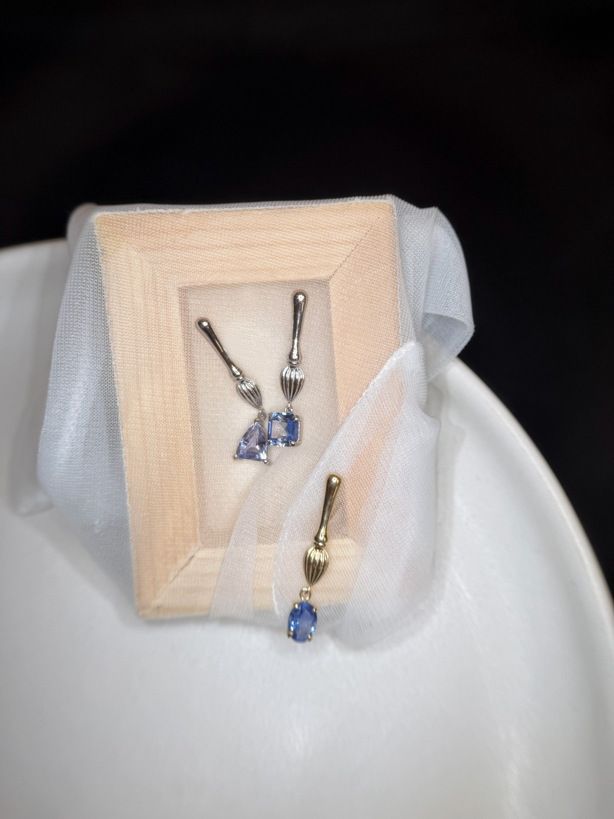Silver and gold drop earrings with blue gemstones displayed on a soft beige stand, gently wrapped in sheer white silk fabric, placed on a white plate.